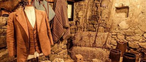 Una habitación rústica de piedra con un maniquí vestido con un abrigo marrón, varias mantas y un interior rústico que incluye paredes de piedra, una ventana y recipientes antiguos.