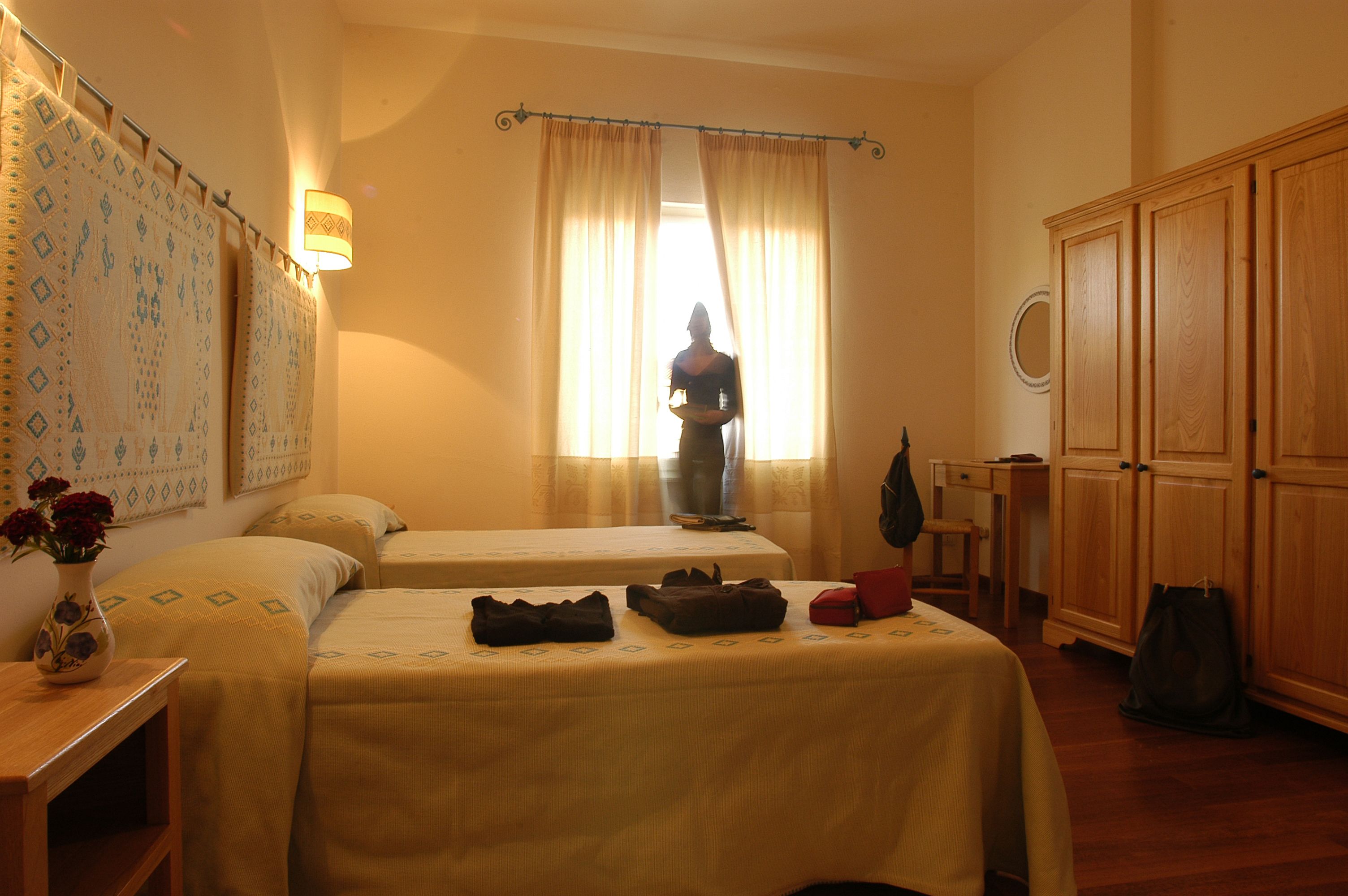 A hotel room with two beds, a nightstand with a vase of flowers, a table with a vase, a suitcase, and a wardrobe.