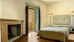 Un dormitorio con suelo de madera, una cama con cubierta color beige, una chimenea y un tapiz de pared estampado.
