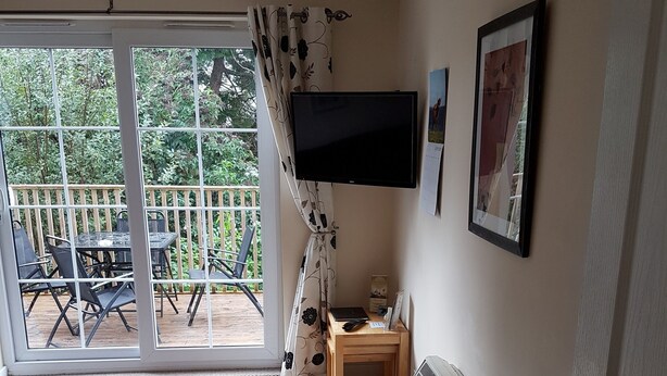 A room with a sliding glass door leading to an outdoor area with a table and chairs. There is a television mounted on the wall, a framed picture, and a small wooden table with a bowl and a remote control. - Apartment 3 Pheonix Flats (Portree)
