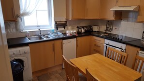 A compact kitchen with wooden cabinets, a built-in dishwasher, a window with curtains, and a wooden dining table. - Apartment 3 Pheonix Flats (Portree)