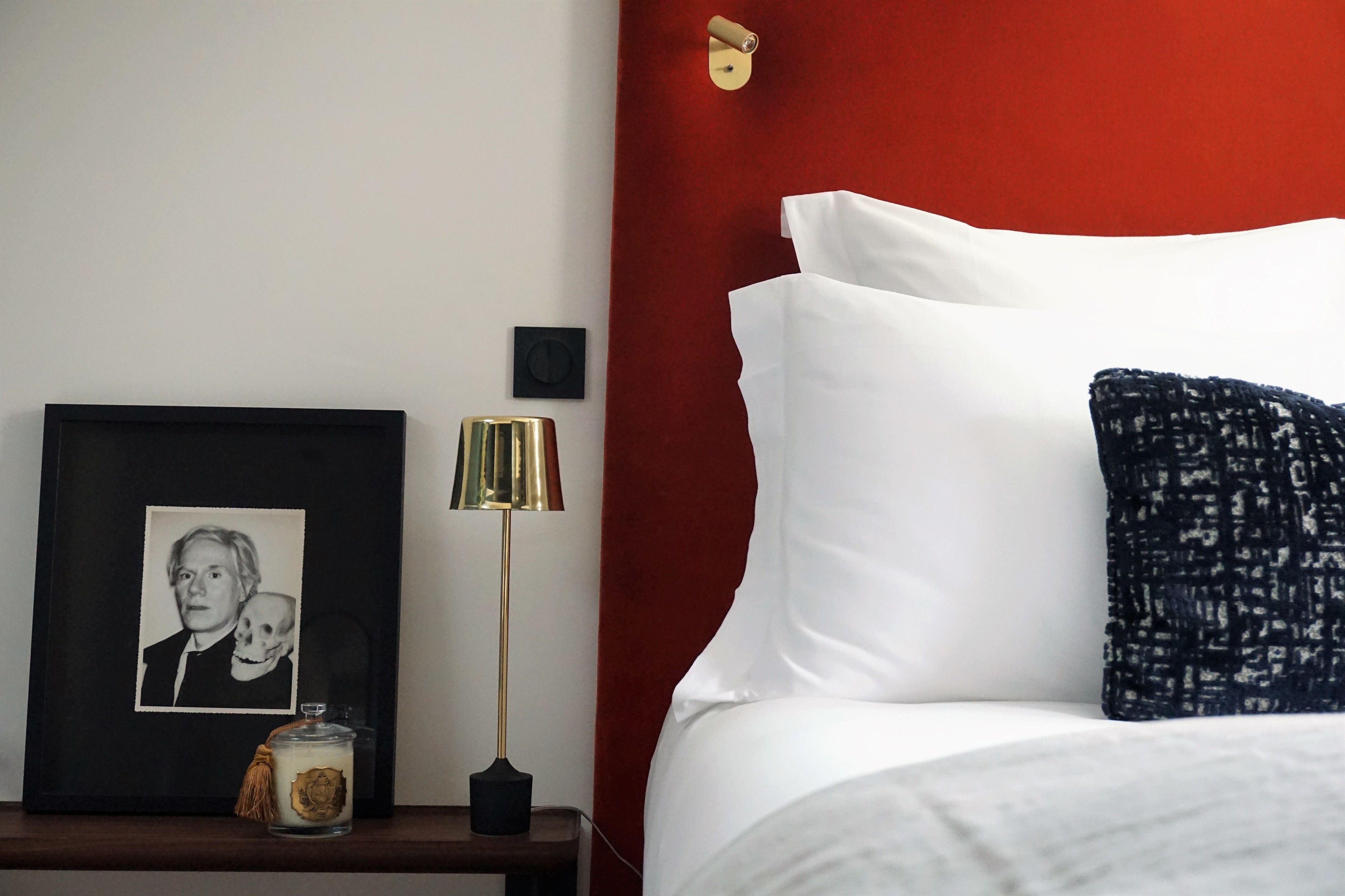 Een net opgemaakt bed met witte en donkerblauwe kussens, een ingelijste foto op een nachtkastje, een lamp en een klein decoratief object.