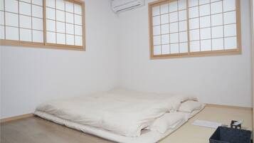 Ein Zimmer mit einem Bett, zwei Fenstern mit Holzrahmen, einem Deckenventilator und einer kleinen schwarzen Aufbewahrungsbox.