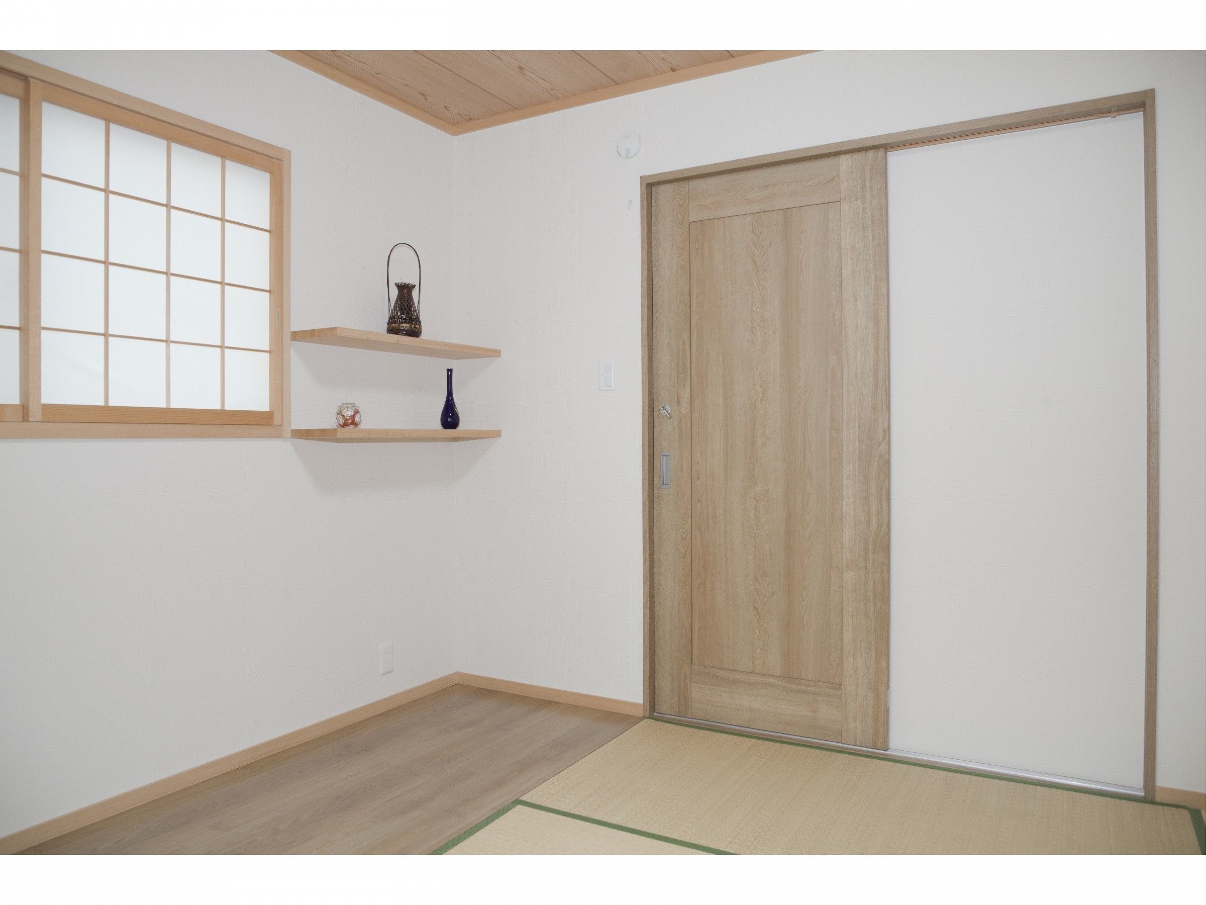 Una habitación con puerta corredera, suelo de madera, ventana con diseño de cuadrícula y dos estantes con adornos.