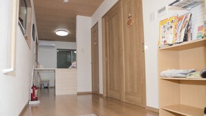 A hallway with wooden flooring, a fire extinguisher, a pair of slippers on a mat, and a shelf with magazines and shoes. - Guest House Aoi Nakamoto (Kyoto)