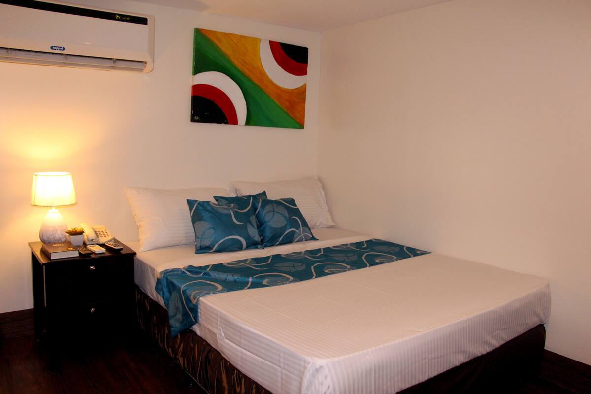 A neatly made bed with a white mattress and blue patterned bedspread, a bedside lamp, a small table with a lamp, a phone, and a book.