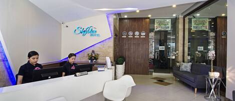 A reception area with a white desk, two receptionists, a reception sign, a clock, and a sofa.
