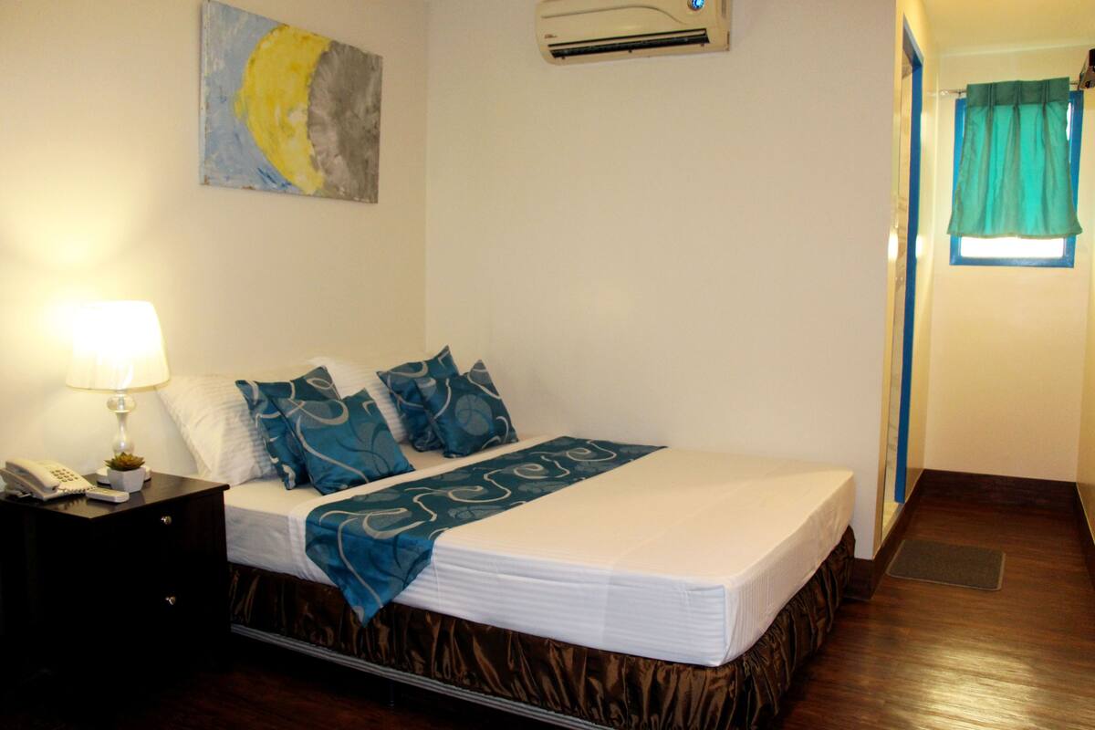 A hotel room with a bed, a nightstand with a lamp, a painting on the wall, and an air conditioning unit.