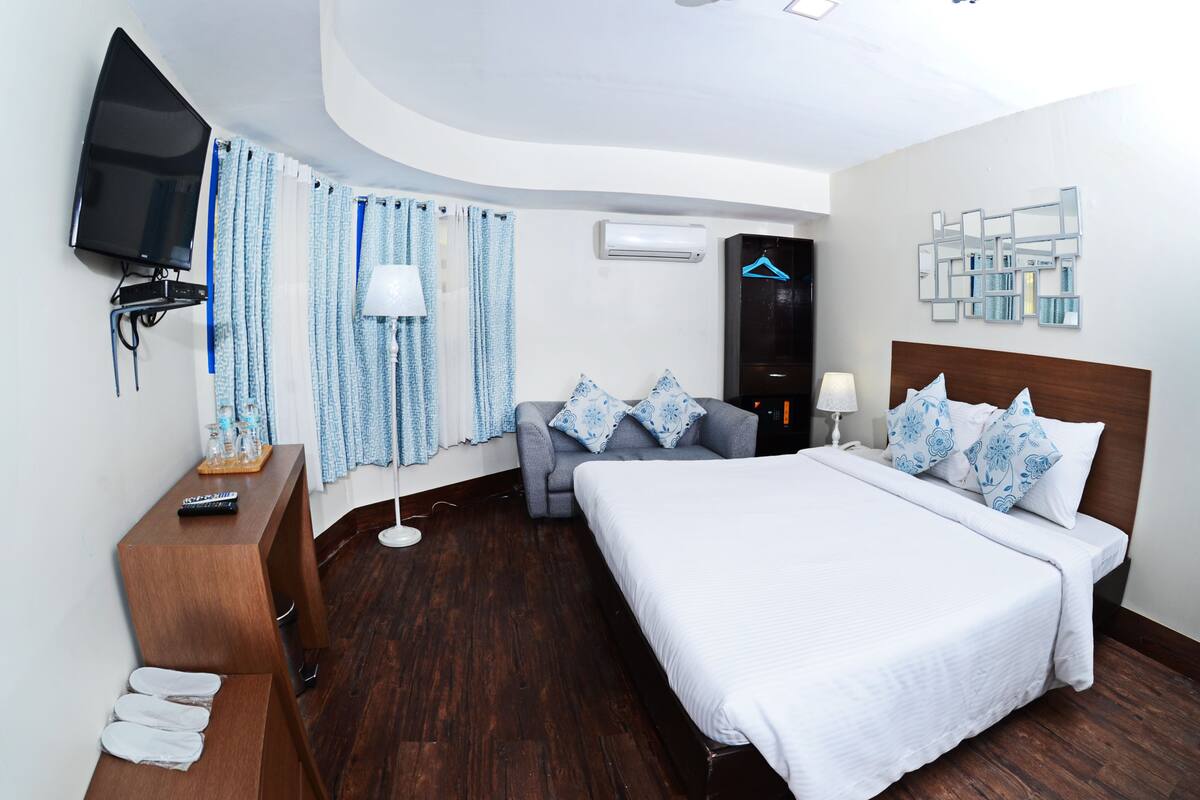A modern hotel room with a large bed, a TV mounted on the wall, a desk with a lamp, and a wooden floor.