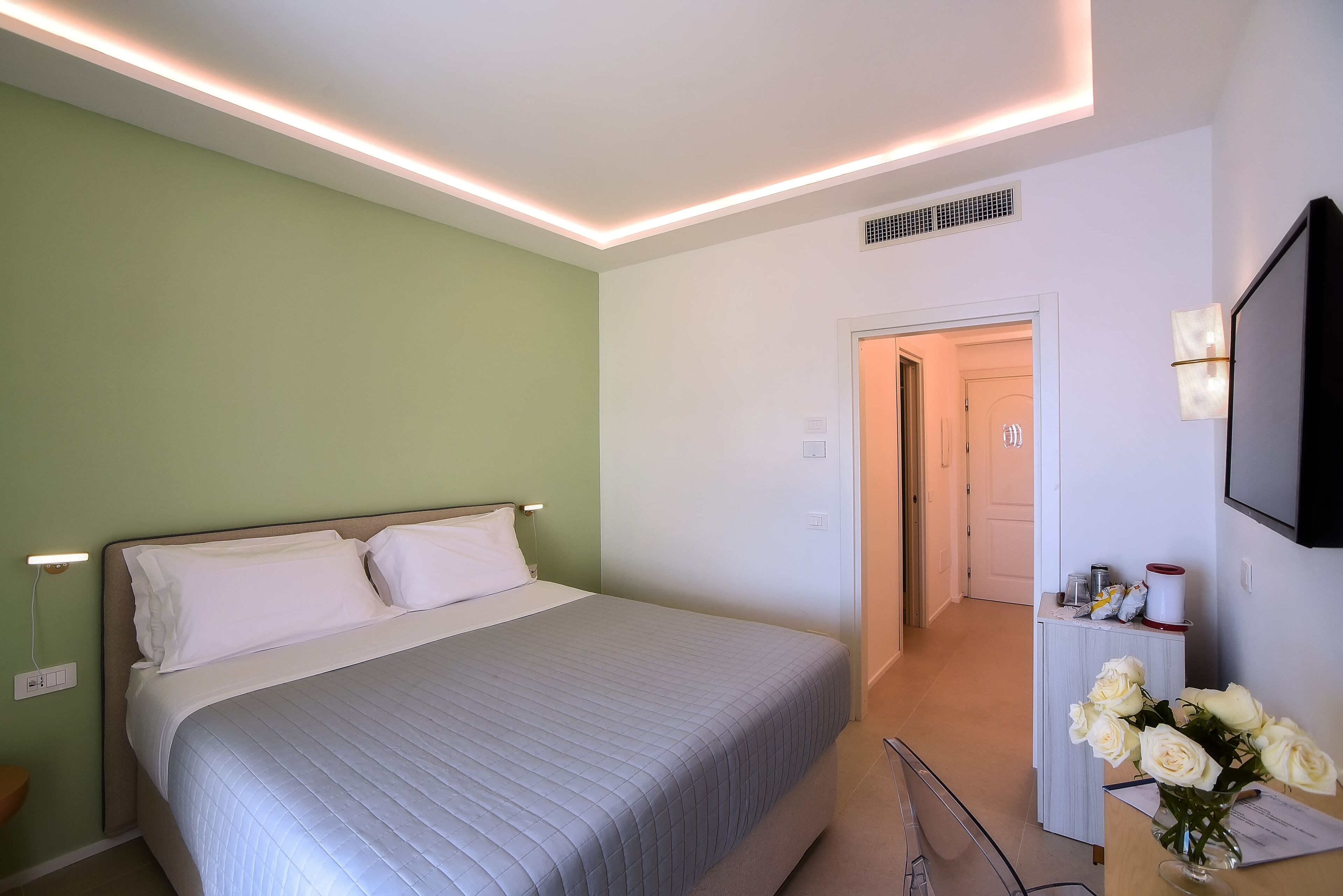 Ein Hotelzimmer mit einem Bett, einem Schreibtisch mit Blumenvase und einem an der Wand befestigten Fernseher.