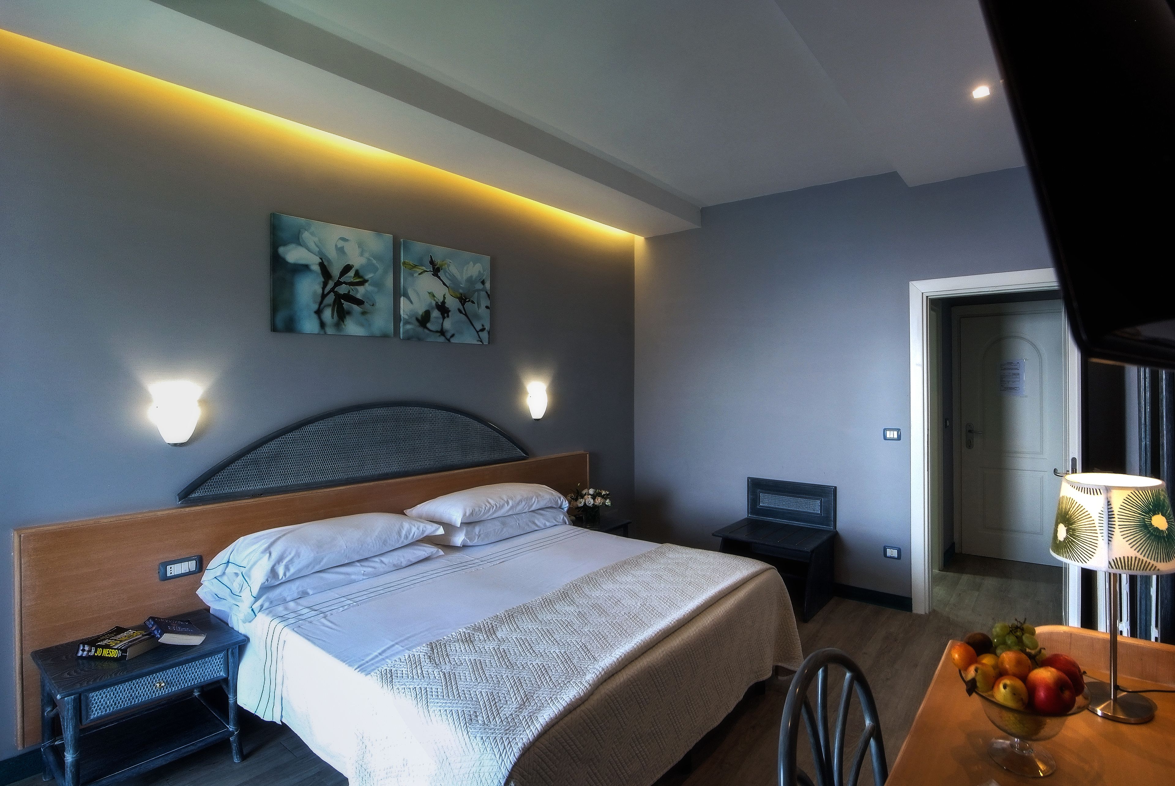 Una habitación de hotel moderna con una cama bien hecha, una mesita de noche pequeña con un libro y un control remoto, un frutero sobre una mesa de madera y un cuadro en la pared.