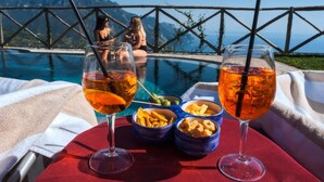 Mesa junto a la piscina con dos vasos de una bebida, cuencos de aperitivos y vistas a un paisaje montañoso.