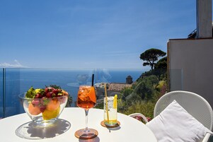A table with two drinks and a fruit bowl, overlooking a scenic view of the sea and hills.