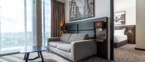 A modern hotel room with a large window, a sofa, a small table, and a bed with a headboard.