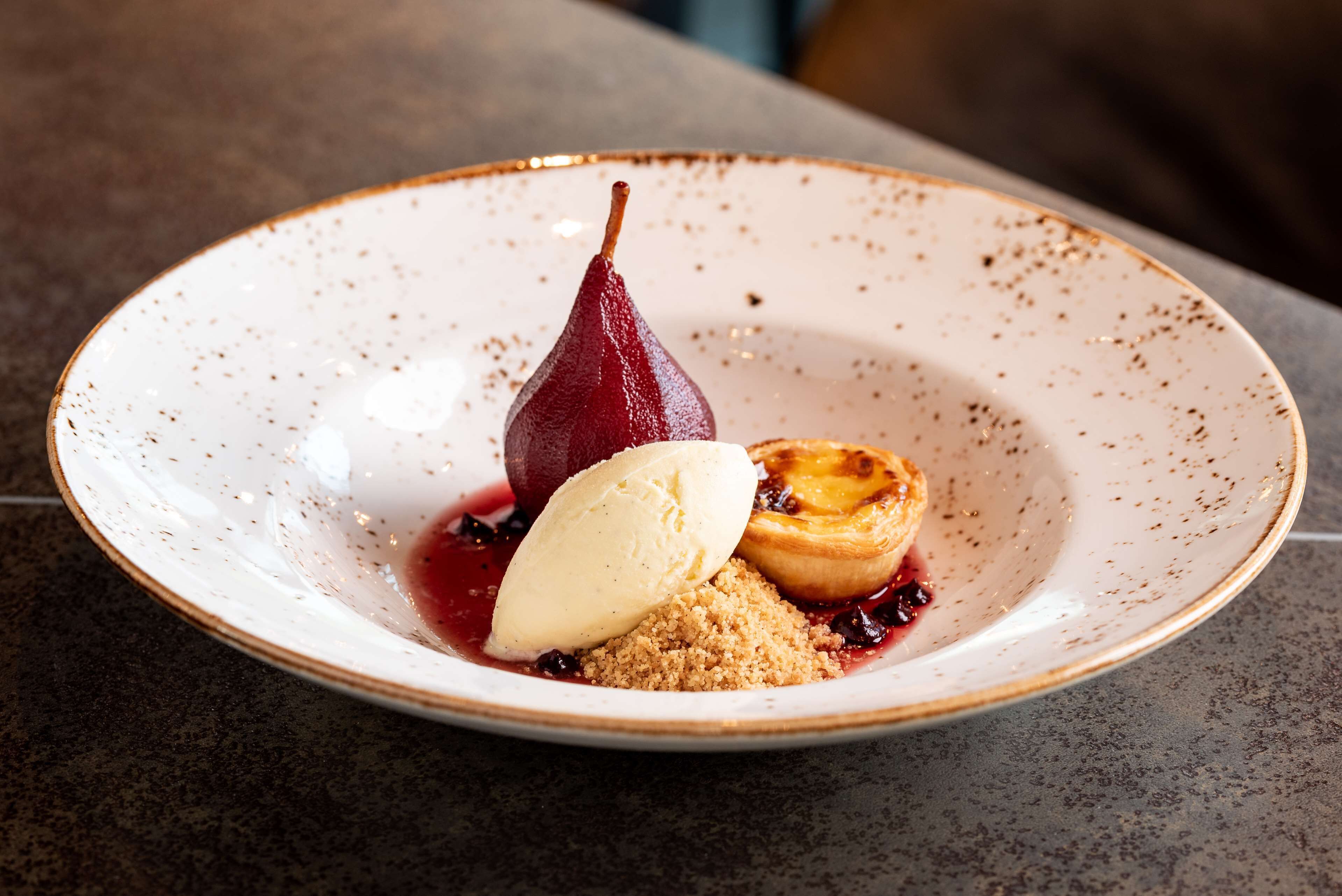 A dessert plate with a pear, ice cream, and a tart on a speckled ceramic plate.