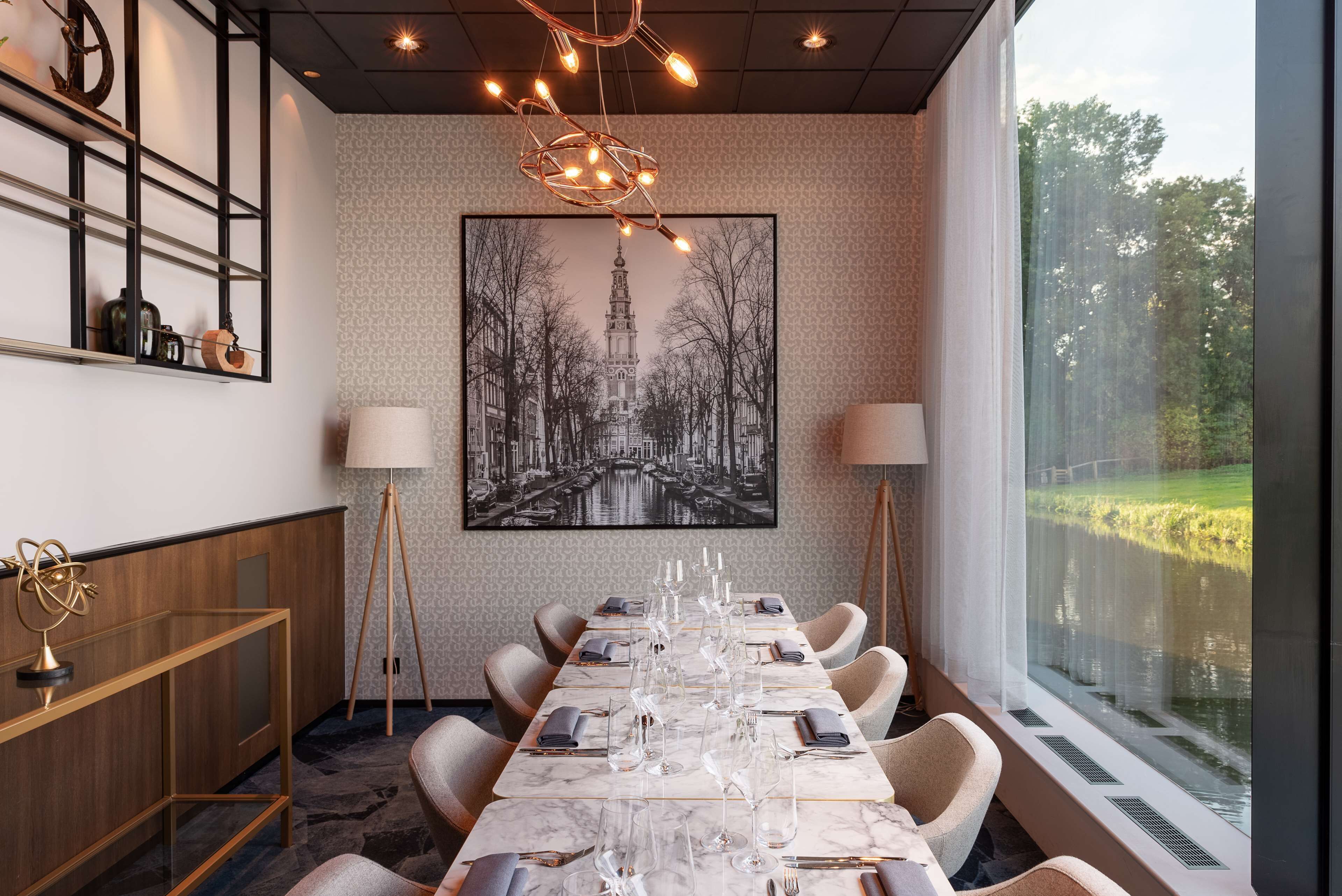 Un espace repas avec une longue table dressée pour six personnes, entourée de chaises, surmontée d’un lustre et agrémentée d’un grand tableau représentant un paysage urbain, encadré.