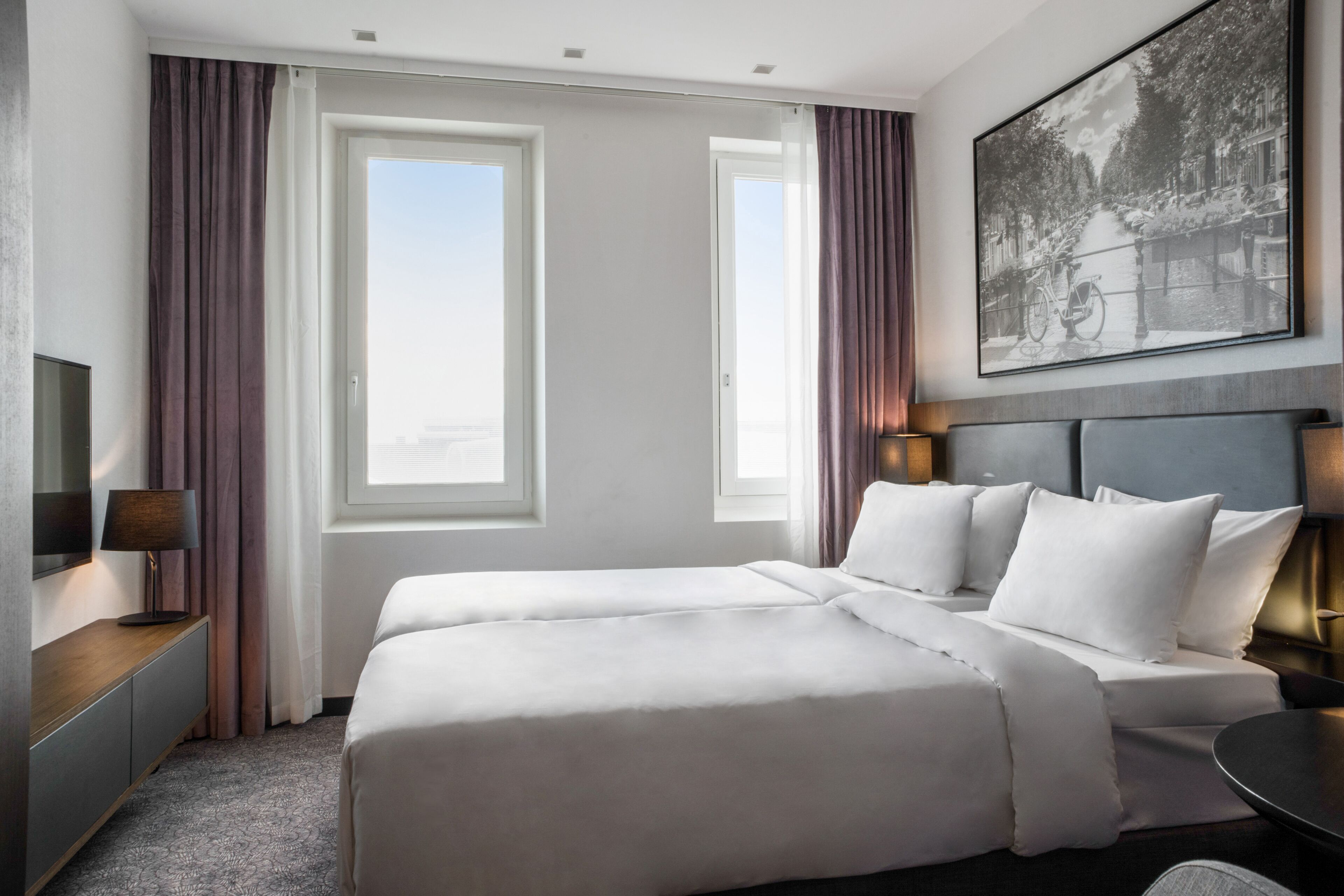 Ein Hotelzimmer mit einem großen Bett, zwei Fenstern, einem Bild an der Wand und einem Schreibtisch mit einer Lampe.