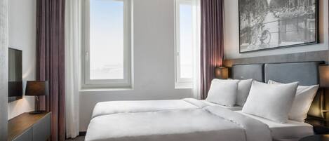 Ein Hotelzimmer mit einem großen Bett, zwei Fenstern, einem Bild an der Wand und einem Schreibtisch mit einer Lampe.