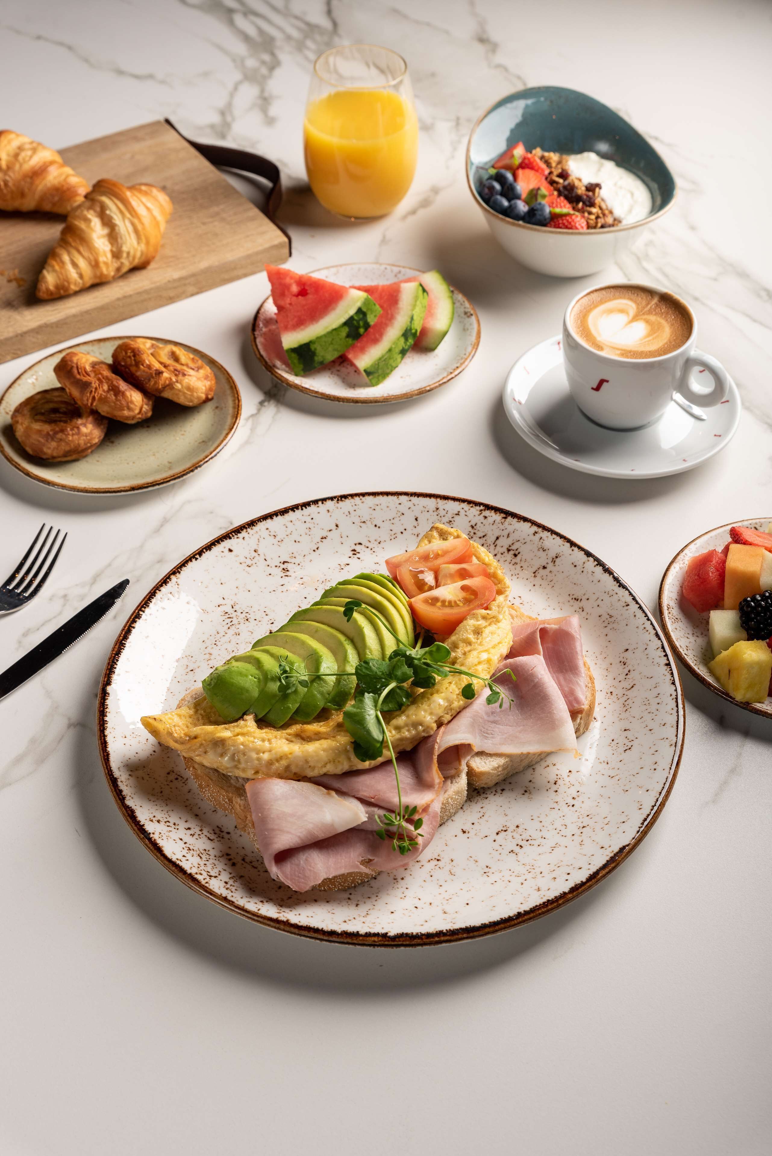 Un desayuno con cruasanes, un tazón de yogur con frutas, un vaso de jugo de naranja, un plato con huevos, aguacate, tomate y jamón, y una taza de café.