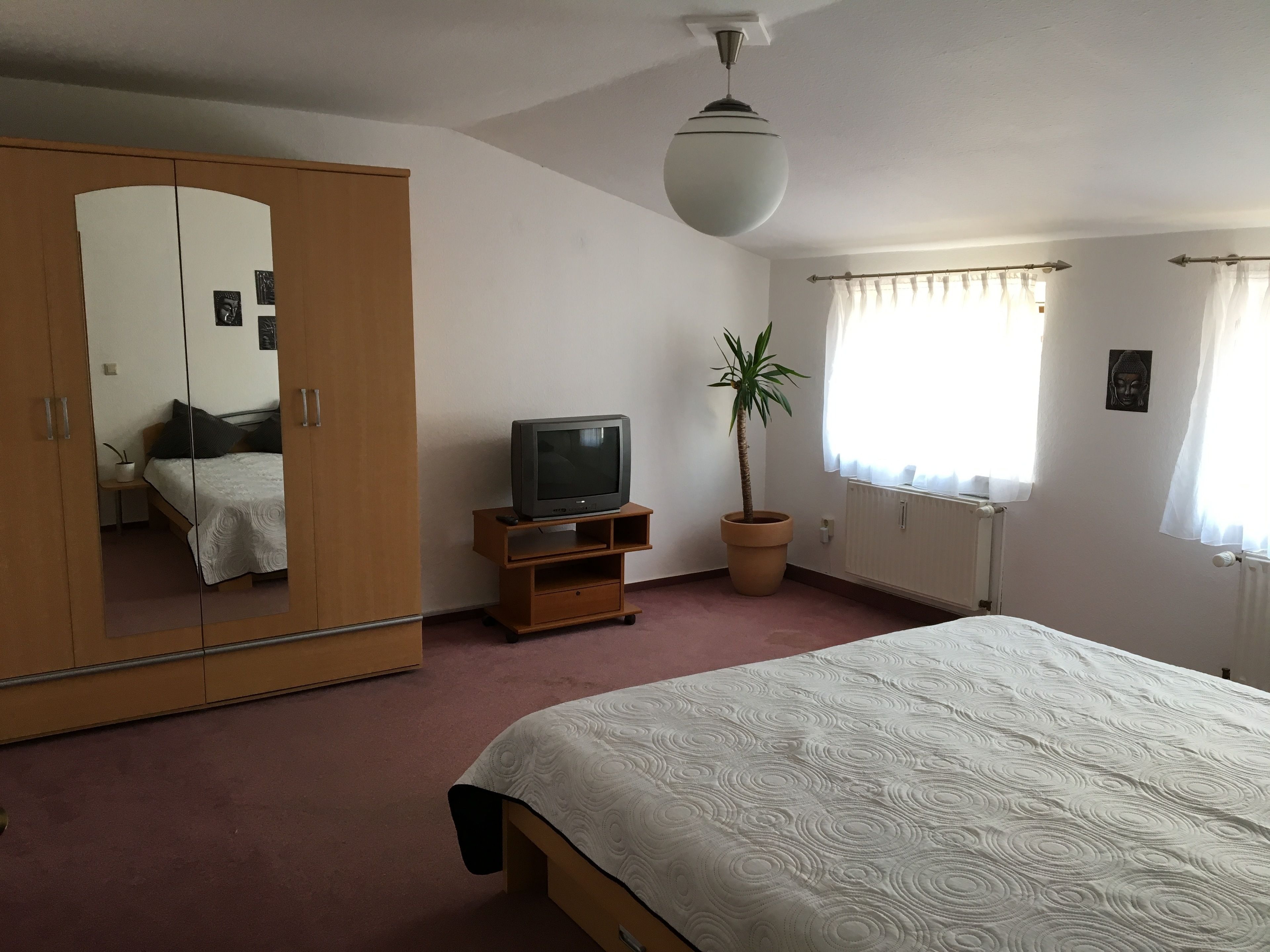 A bedroom with a bed, a wardrobe, a television on a wooden stand, a potted plant, and a window with curtains.