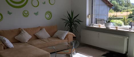 Un salon moderne avec un canapé beige, une table basse en verre, un tapis en gazon synthétique vert et une fenêtre donnant sur un paysage verdoyant.