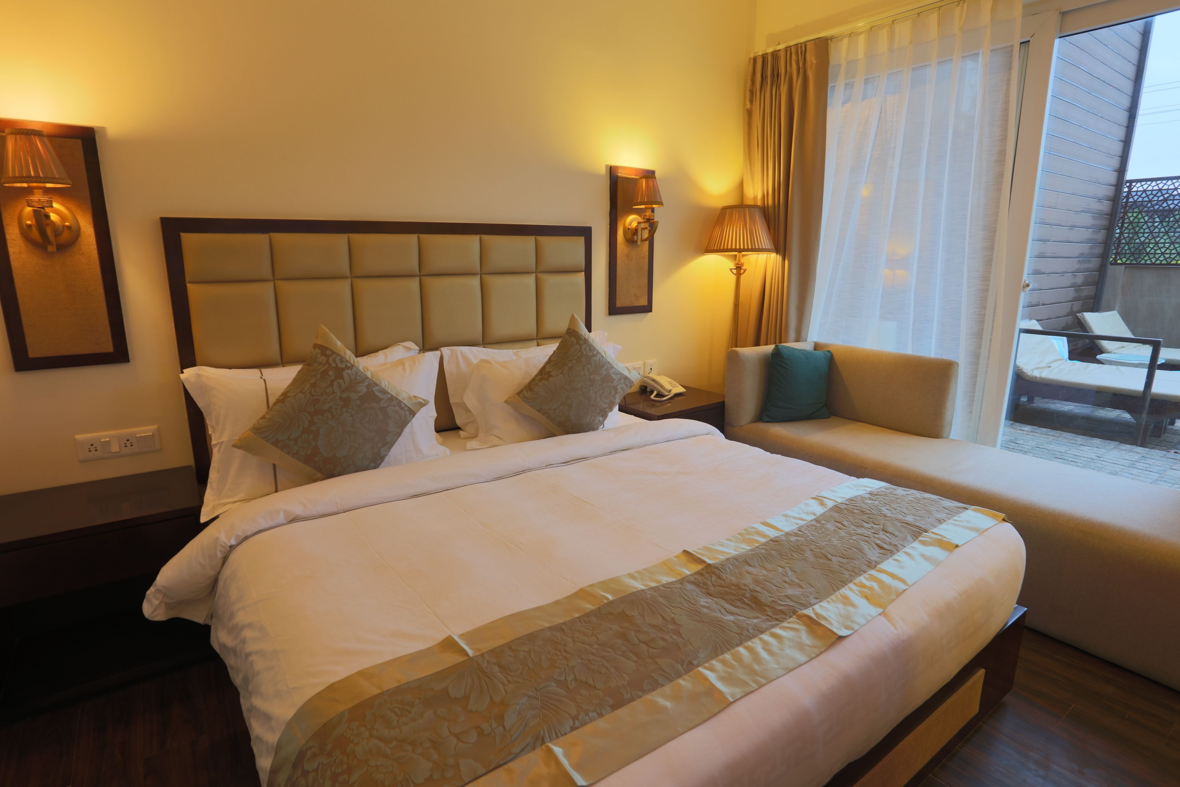 Quarto de hotel com uma cama grande, um sofá, varanda com vista e duas luminárias de parede.