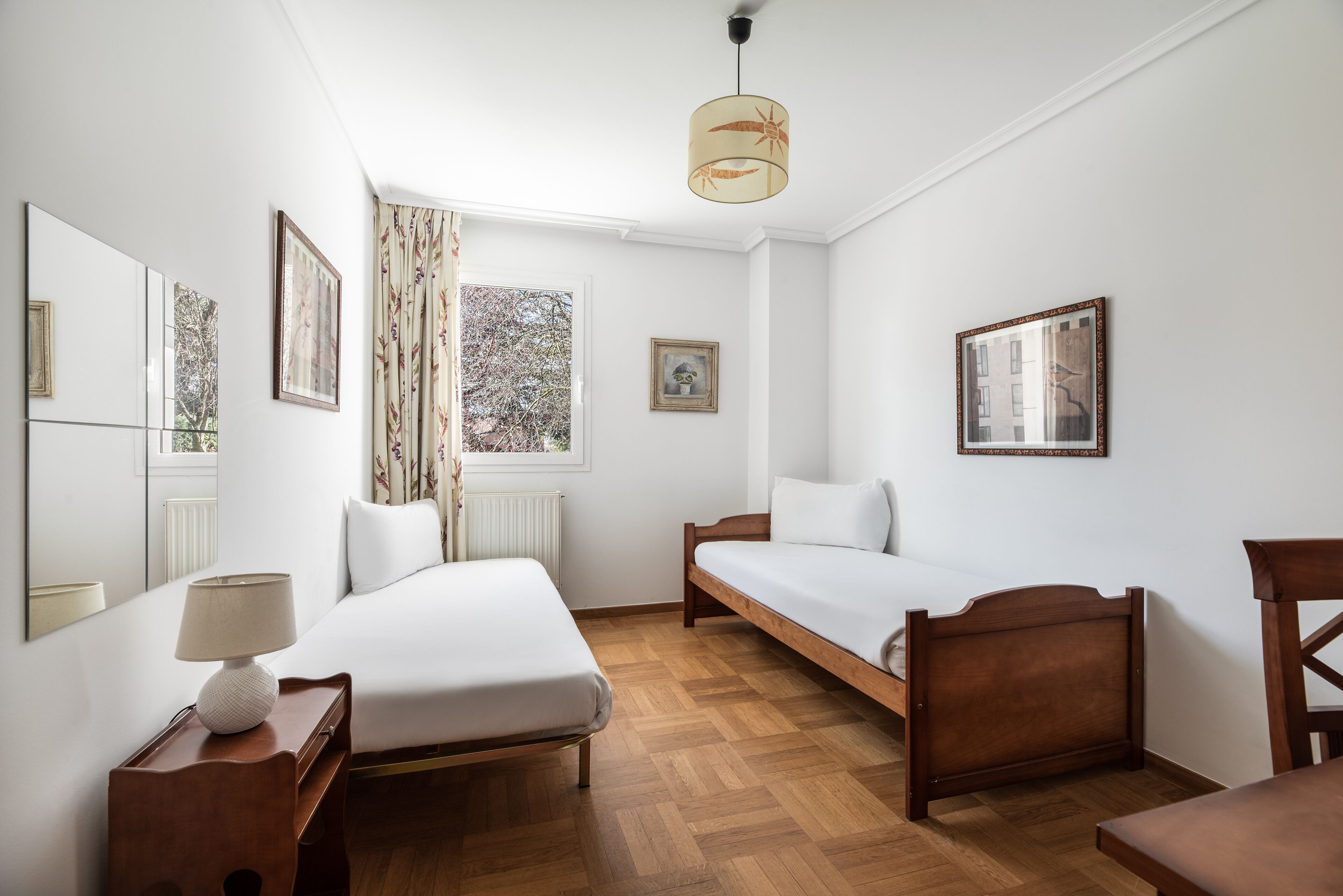 A room with two beds, a wooden floor, a window with curtains, and a ceiling lamp.