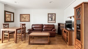 A living room with a brown leather sofa, wooden coffee table, and matching chairs. There is a TV on a wooden stand, a cabinet with glass doors, and framed artwork on the walls. - Apartamentos El Cid (Burgos)