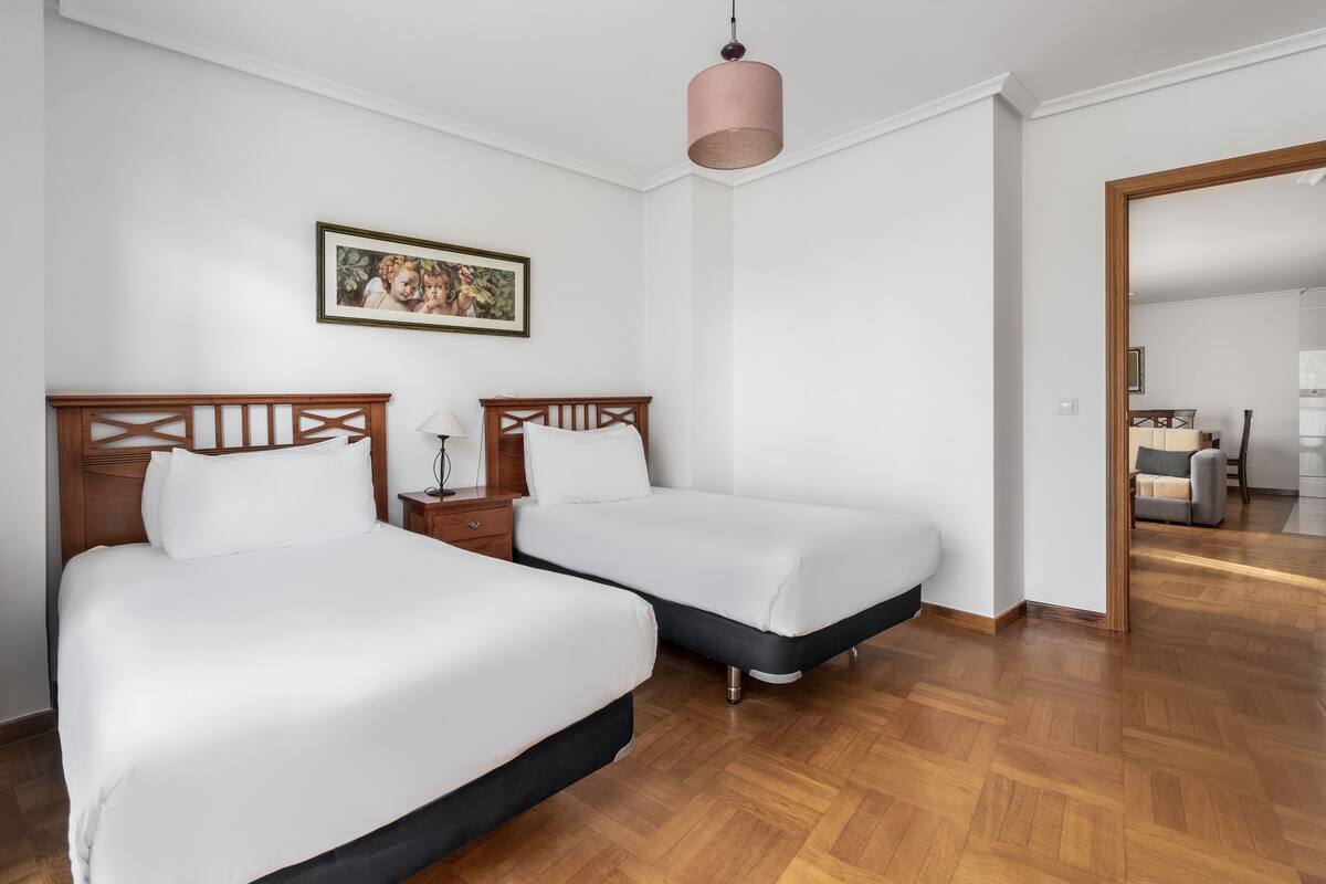 A hotel room with two beds, a wooden headboard, a bedside table, a lamp, a framed picture on the wall, and a mirror in the corner.