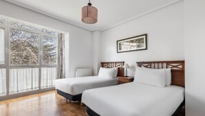 A hotel room with two beds, a wooden headboard, a bedside table with a lamp, a window with curtains, and a framed picture on the wall. - Apartamentos El Cid (Burgos)