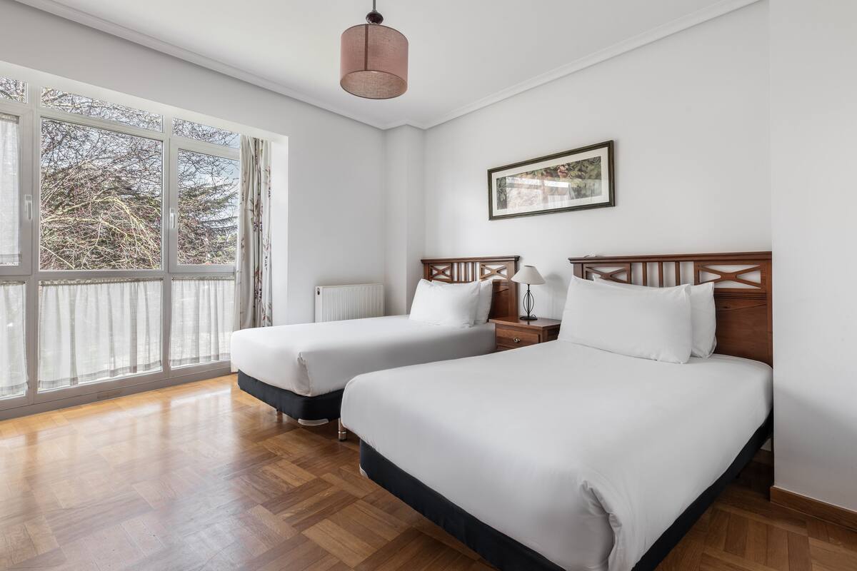 A hotel room with two beds, a wooden headboard, a bedside table with a lamp, a window with curtains, and a framed picture on the wall.