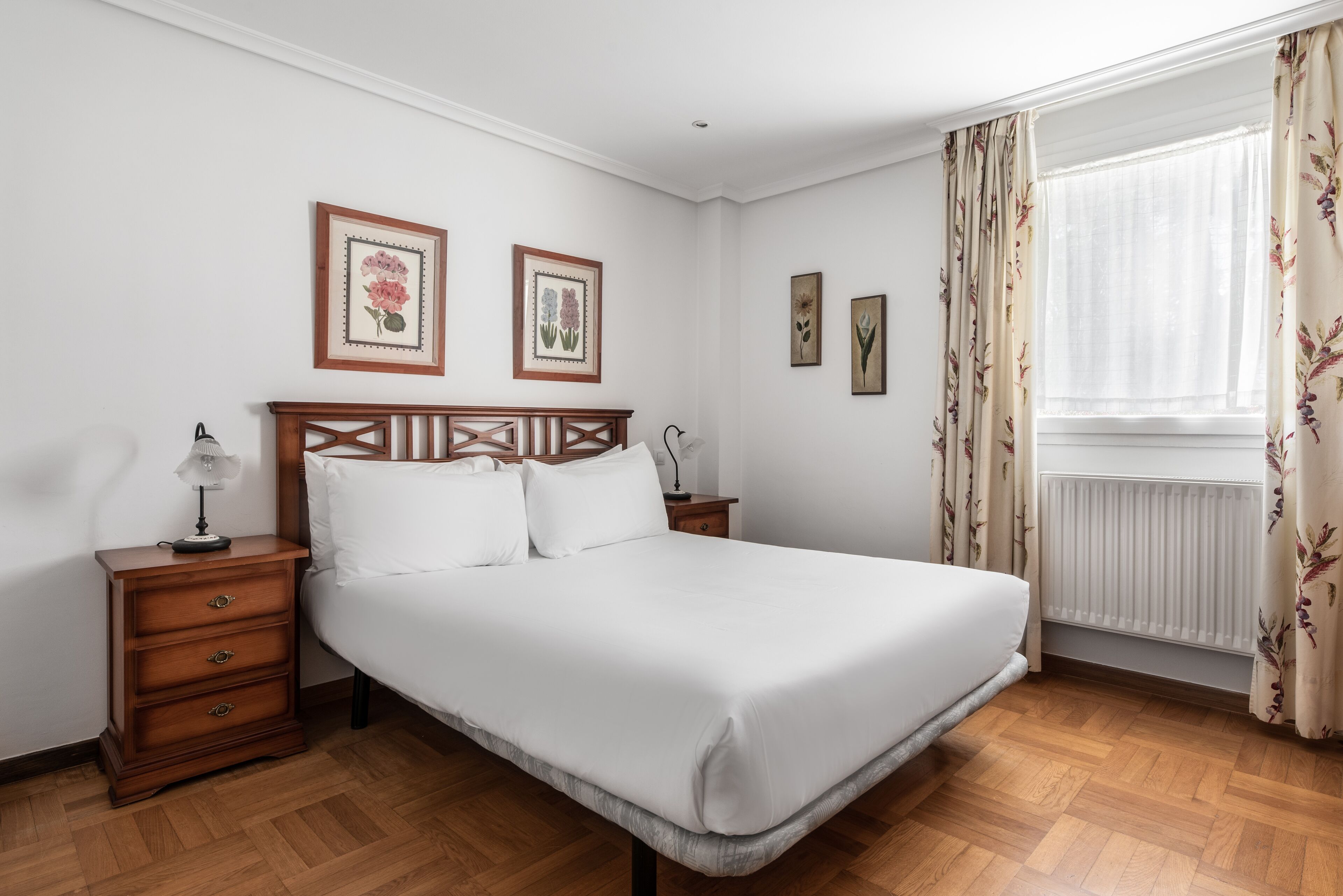 A neatly arranged bedroom with a wooden bed, a nightstand with a lamp, and framed floral artwork on the wall.