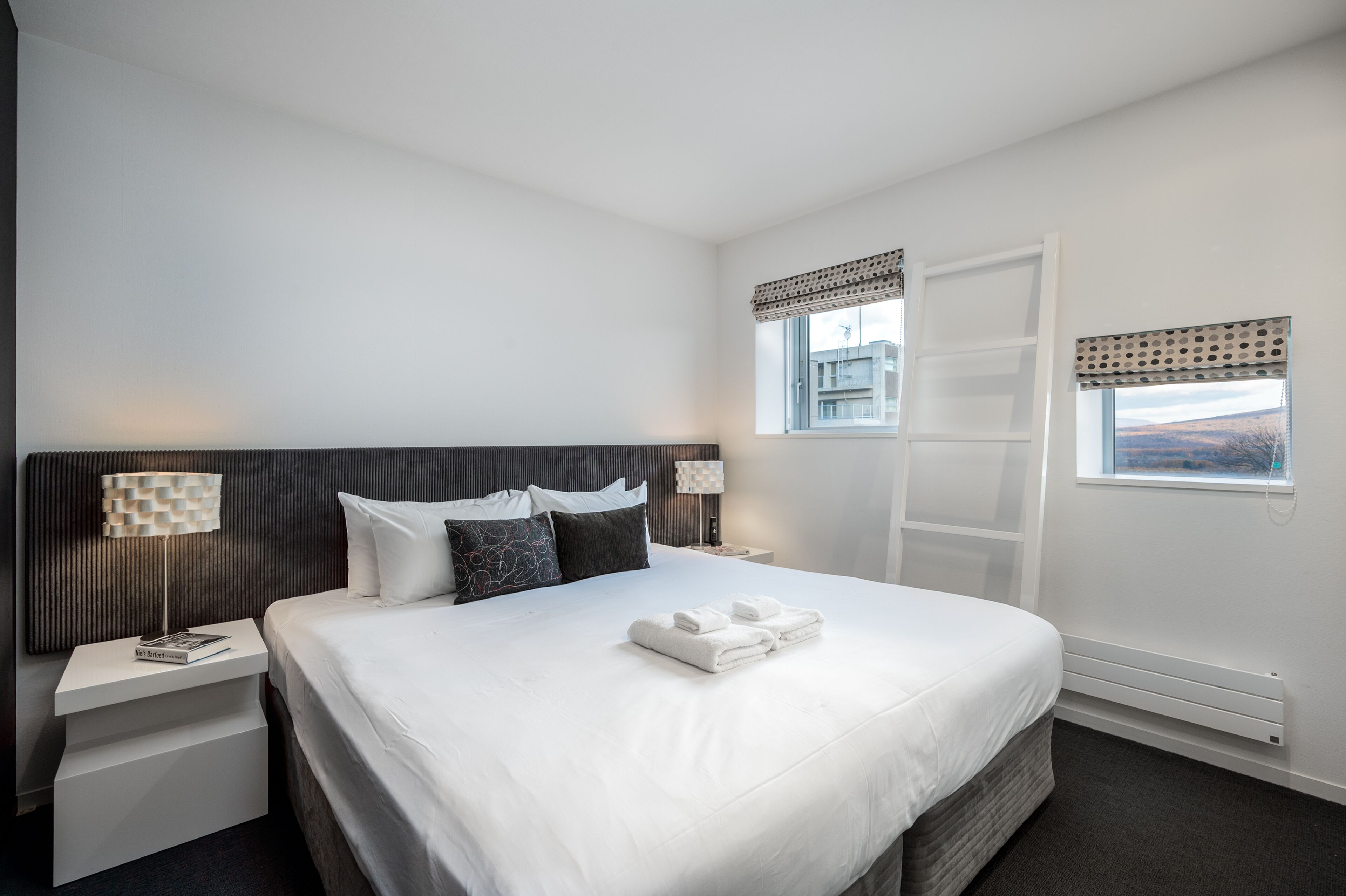 A modern hotel room with a large bed, white linens, a bedside table with a lamp, and a view of the outside through a window.