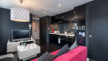 A modern living room with a flat-screen TV, white coffee table, and a red sofa. A kitchen area is visible with a microwave, stove, and cabinets.