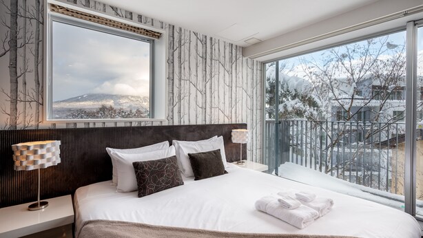 A modern hotel room with a large bed, a view of a snowy landscape, and a balcony with a railing. - Kira Kira Suites by H2 Life (Kutchan)
