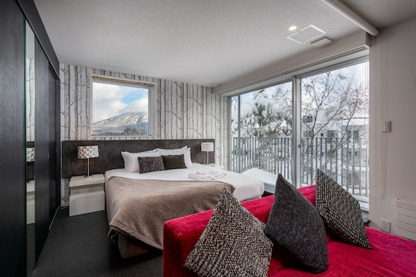 Quarto de hotel moderno com uma cama grande, um sofá vermelho e vista para uma montanha nevada.