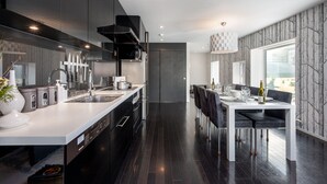 A modern kitchen with dark cabinetry, a white countertop, and stainless steel appliances. There is a dining area with a white table and black chairs. - Kira Kira Suites by H2 Life (Kutchan)