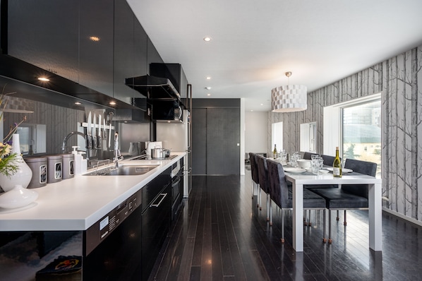 Een moderne keuken met donkere kastjes, een wit aanrechtblad en roestvrijstalen apparatuur. Er is een eethoek met een witte tafel en zwarte stoelen.