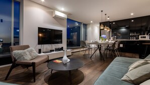 A modern living room with a sofa, a chair, a coffee table, and a TV. There is a dining area with a table and chairs. The kitchen is visible with dark cabinets and a microwave. - Kira Kira Suites by H2 Life (Kutchan)