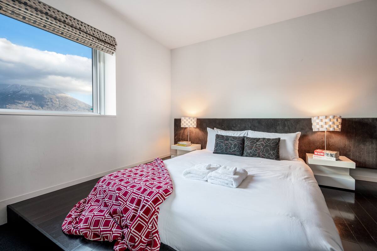 Une chambre d’hôtel moderne avec un grand lit, deux tables de chevet, une fenêtre donnant sur les montagnes et un plaid à motifs.