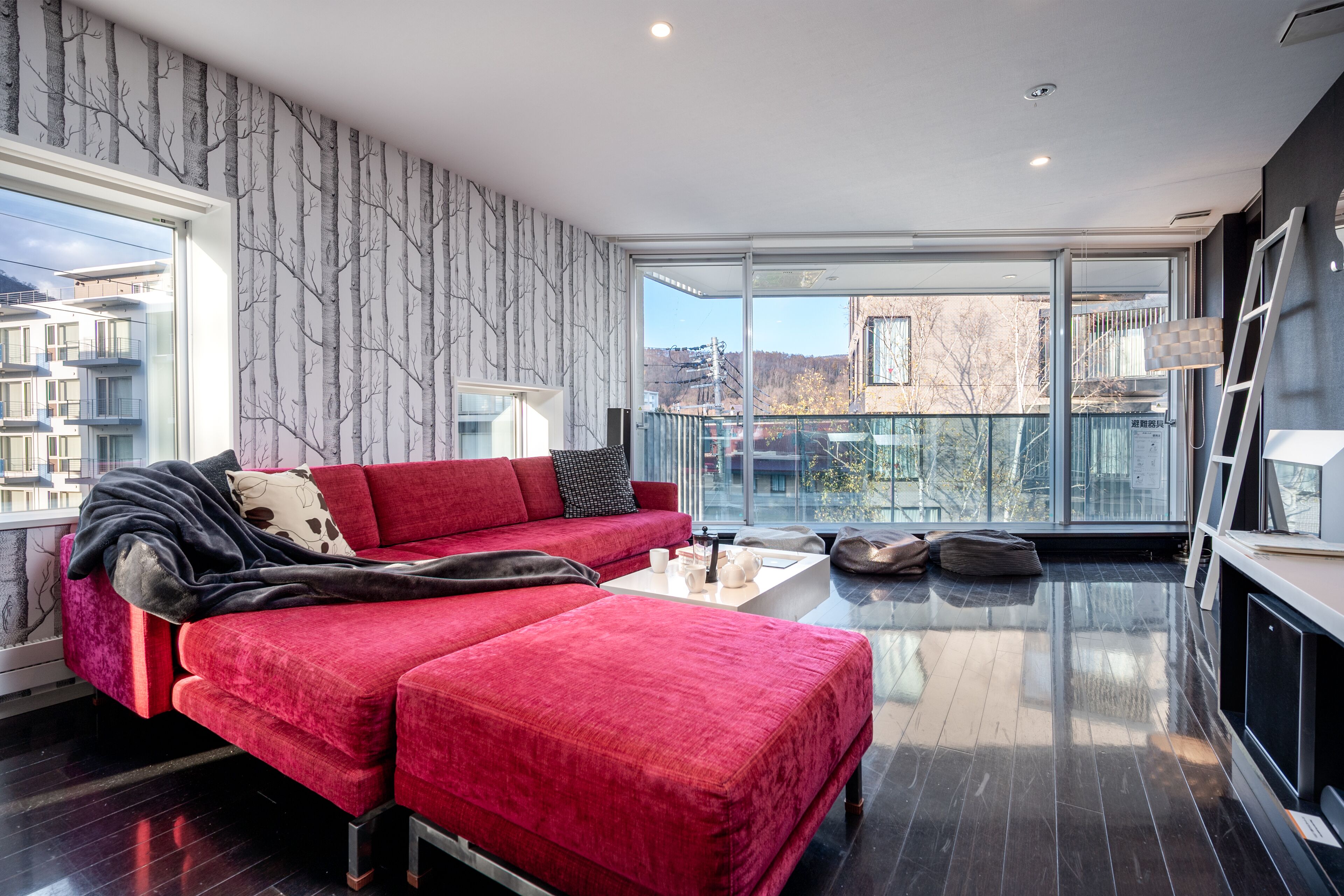 A modern living room with a red sofa, a coffee table, and a view of buildings through large windows.