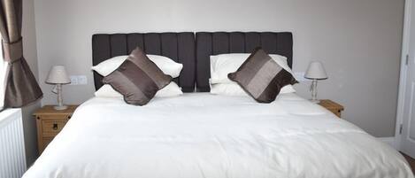 A neatly made bed with white bedding and two dark pillows, a nightstand with a lamp, and a cityscape painting on the wall.
