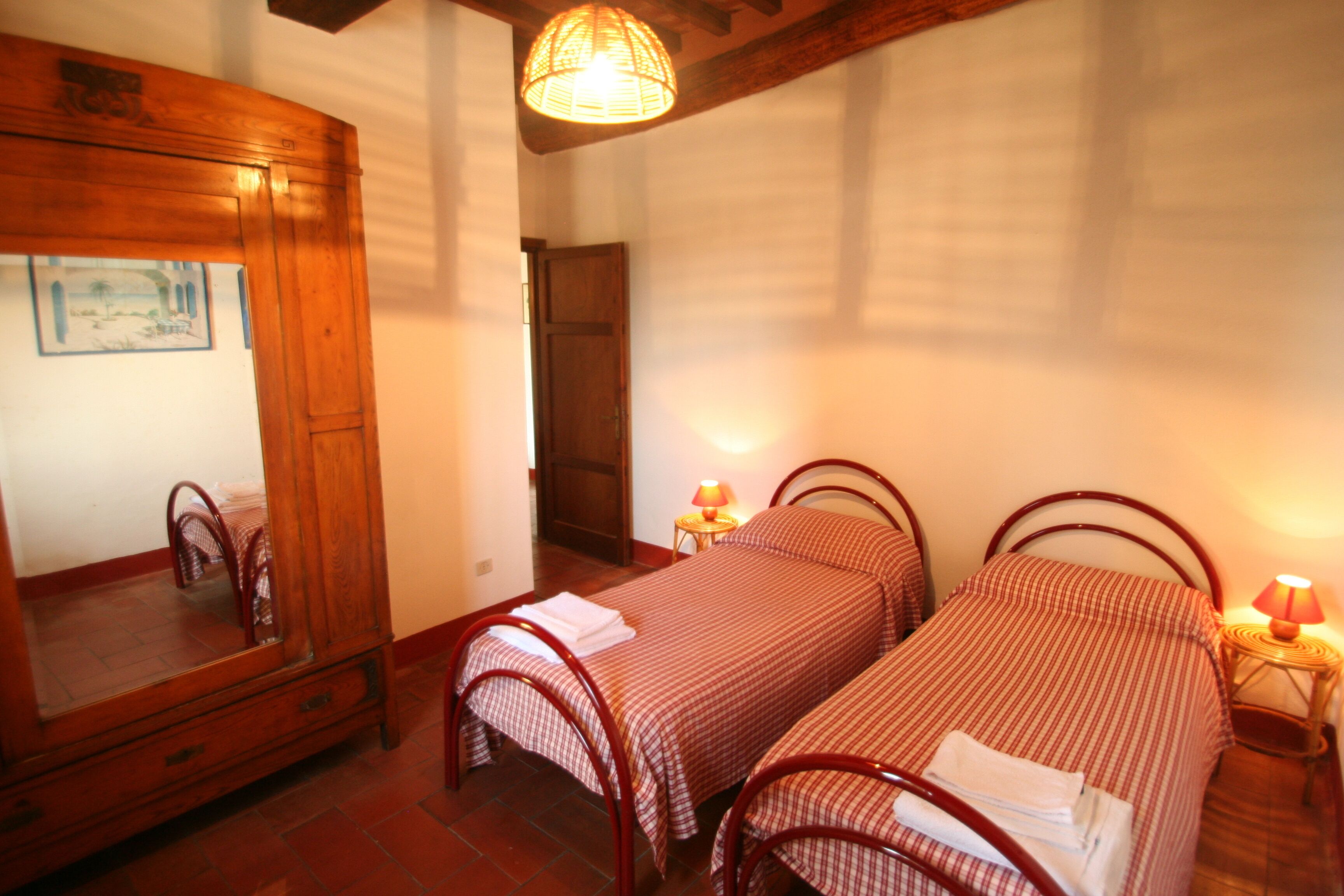 A bedroom with two single beds, a wooden wardrobe, a mirror, and a small table with a lamp.