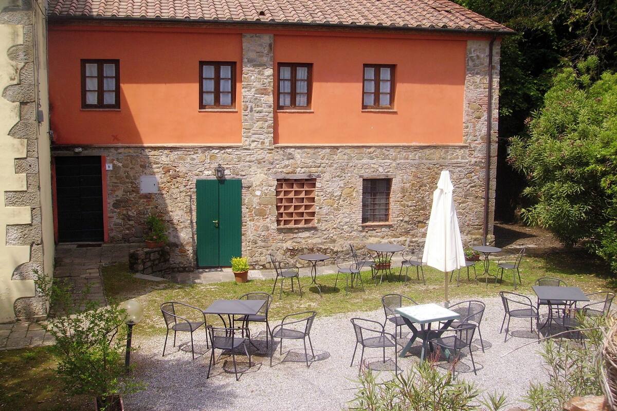 A building with a stone facade, multiple windows, and a green door. There are outdoor tables and chairs arranged in a courtyard.