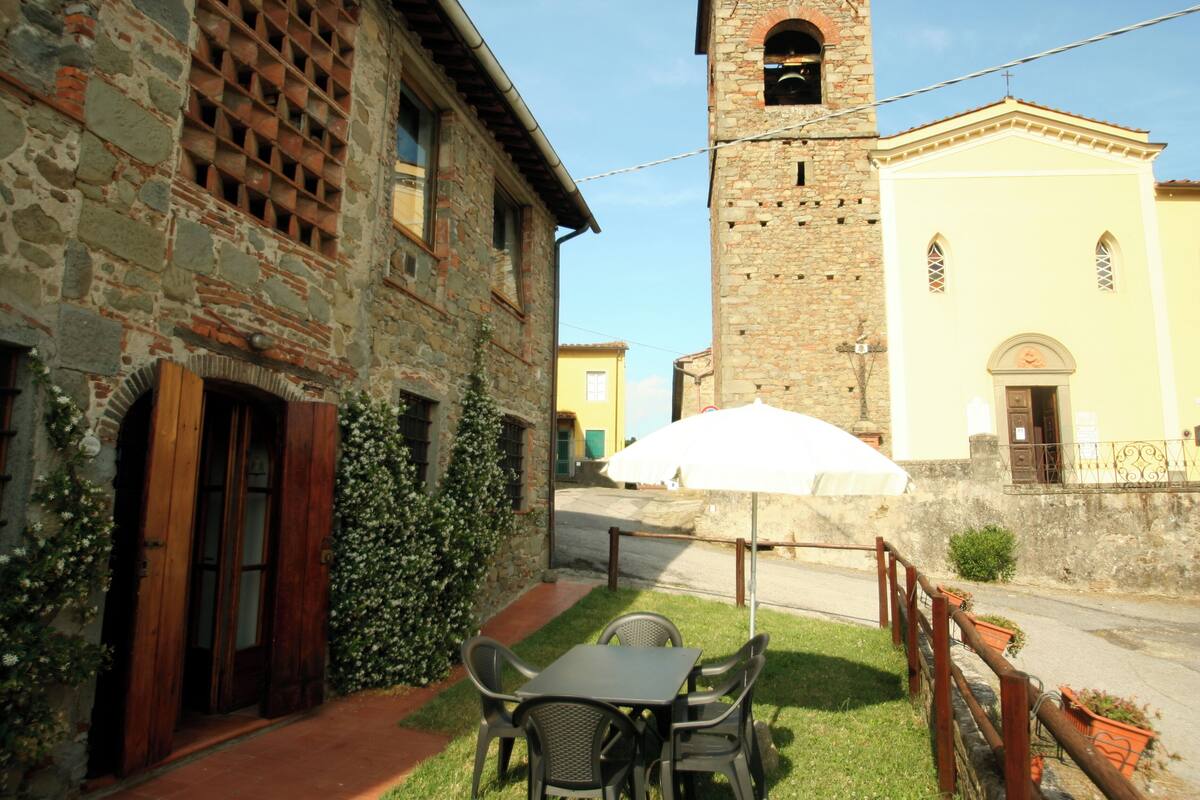 An outdoor seating area with a table and chairs, a white umbrella, and a stone building with a tower in the background.