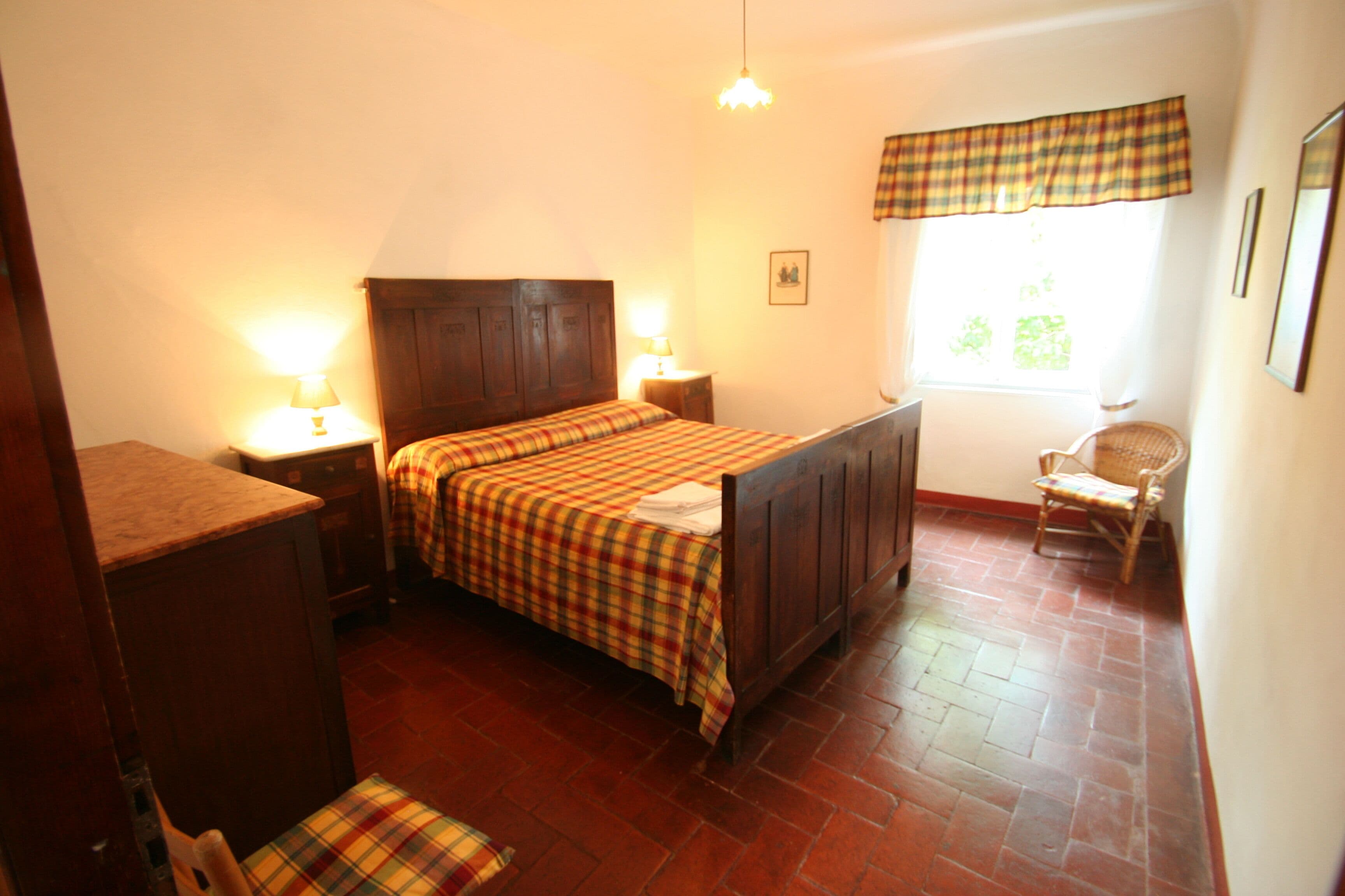 Une chambre à coucher avec un lit en bois, deux tables de chevet avec des lampes, une chaise et une fenêtre donnant sur un paysage verdoyant.