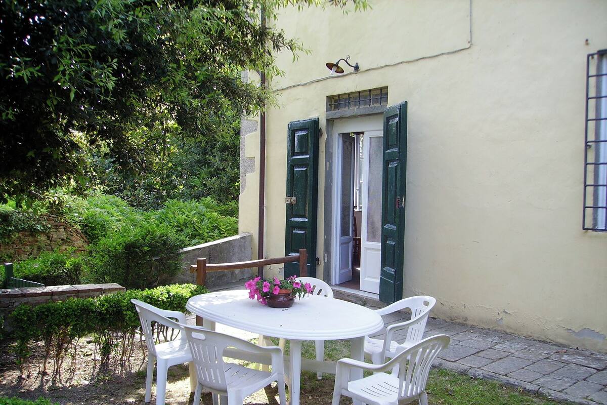 A round white table with four white plastic chairs, a potted plant with pink flowers, and a door with green shutters.