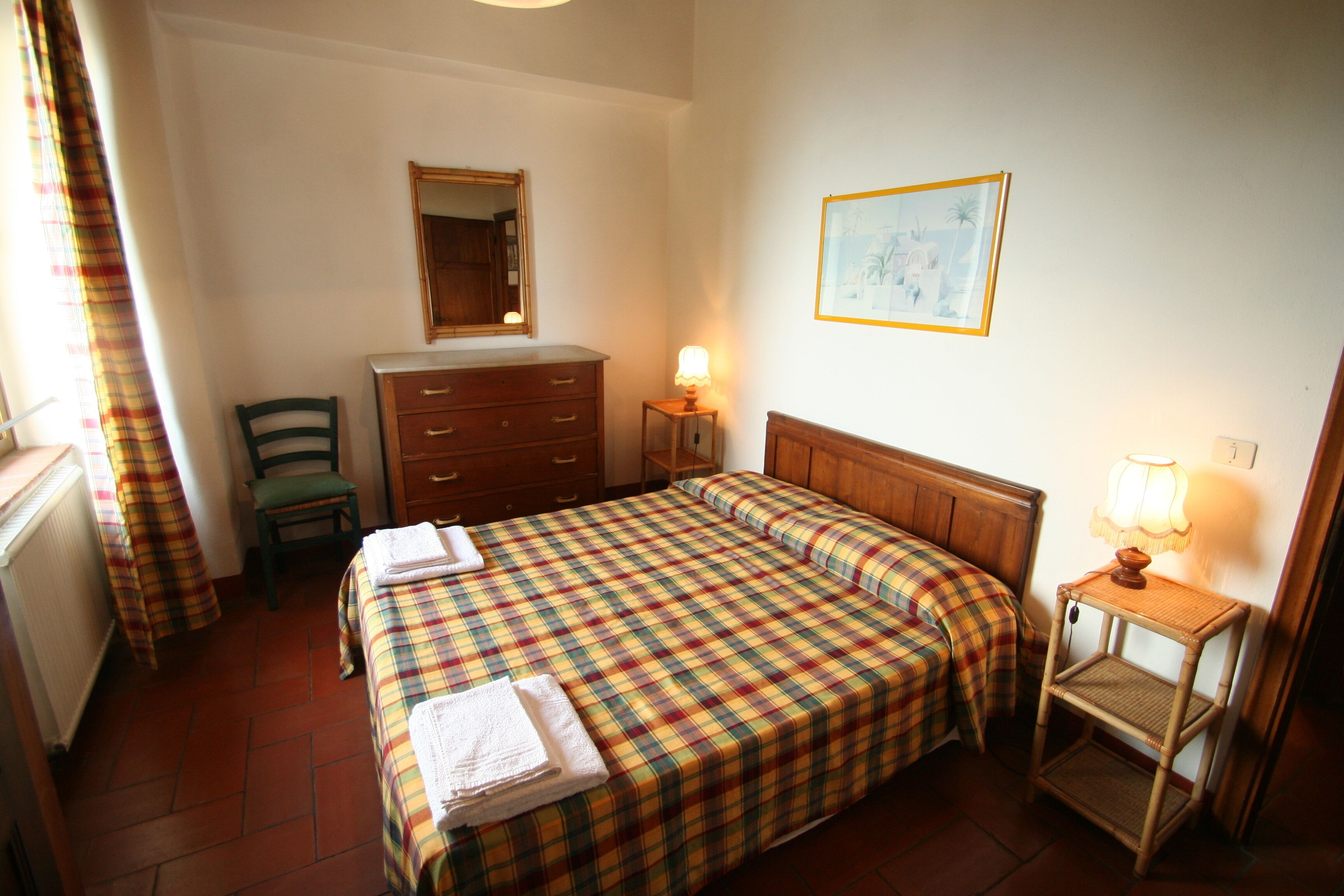 A bedroom with a bed, a dresser, a chair, a bedside table with a lamp, a mirror, and a framed picture on the wall.