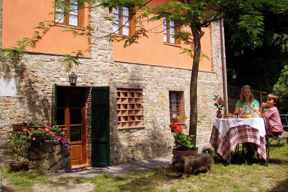 A rustic stone building with a wooden door and shutters, a small table with a checkered tablecloth, and a dog nearby.