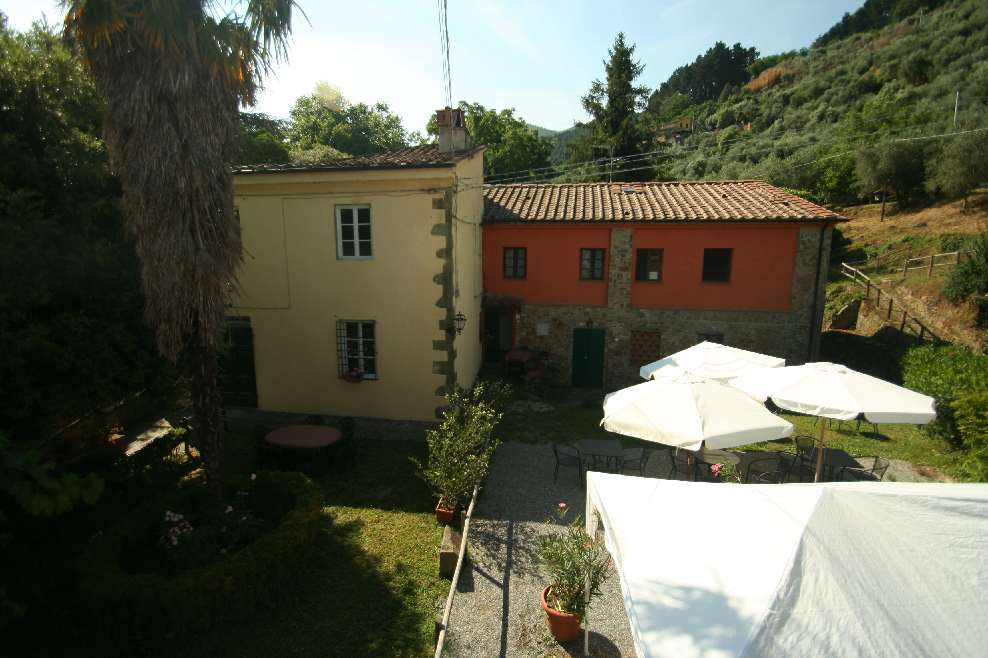 Un bâtiment à deux étages avec une façade jaune et rouge, des tables à l’extérieur sous des parasols, et un espace jardin agrémenté de plantes en pot.